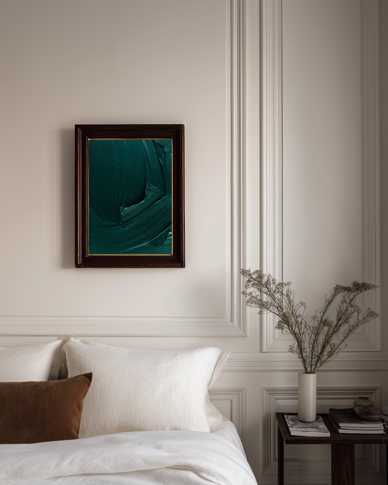 Framed abstract emerald green art on a wall above a bed with a vase of dried flowers on a nightstand.
