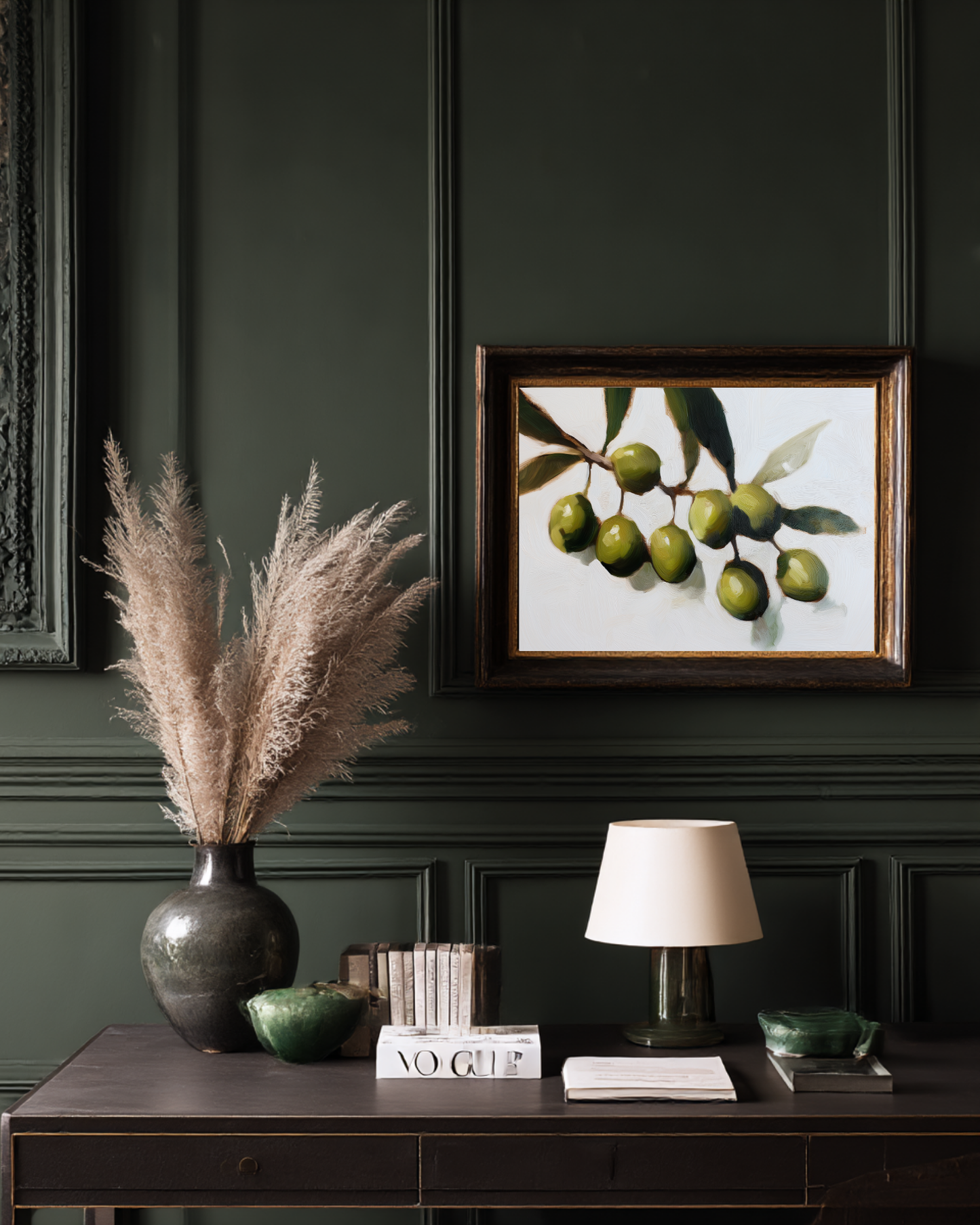 Decorative setup with a vase of dried plants, framed artwork of green olives, and books on a dark surface against a dark wall.