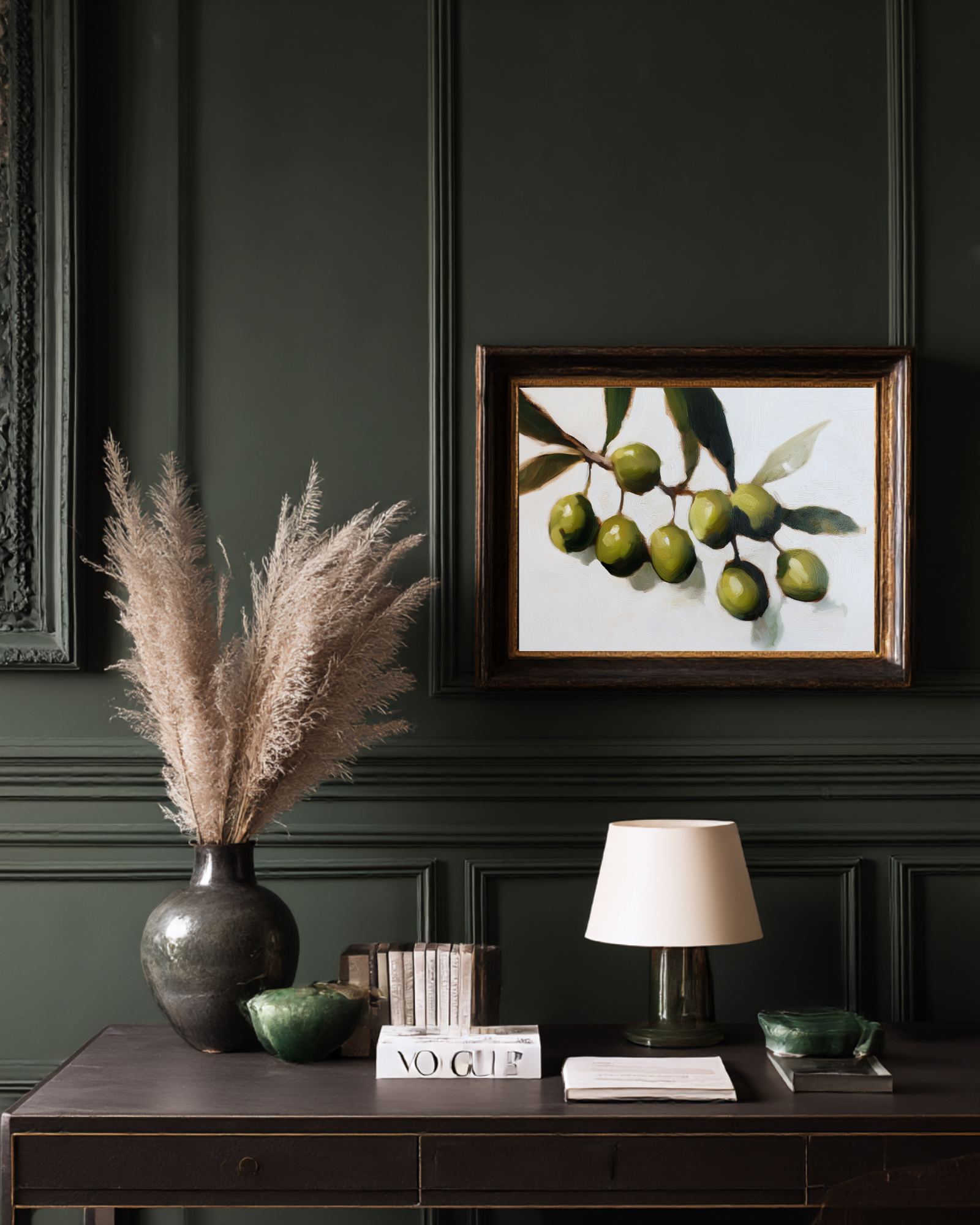 Decorative setup with a vase of dried plants, framed artwork of green olives, and books on a dark surface against a dark wall.