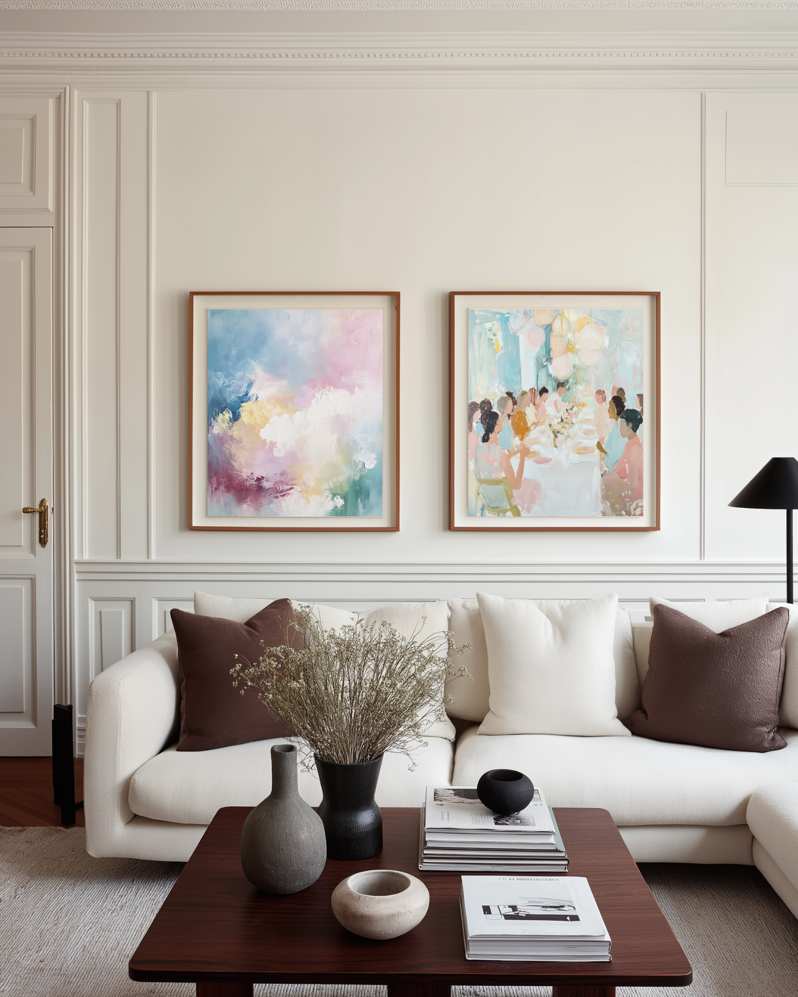 Living room with a white sofa, decorative pillows in white and dark brown, and a dark brown coffee table with books and a vase. Two pastel watercolor paintings on a French white wall. 